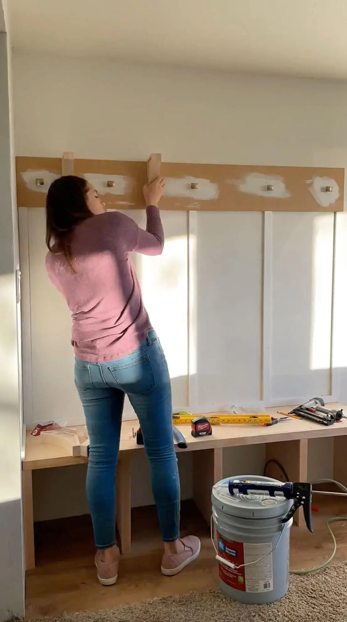 A woman installing decorative support brackets beneath a wooden shelf above a built-in bench. The project is coming together with a combination of storage cubbies, board and batten wall design, and sturdy wooden construction.